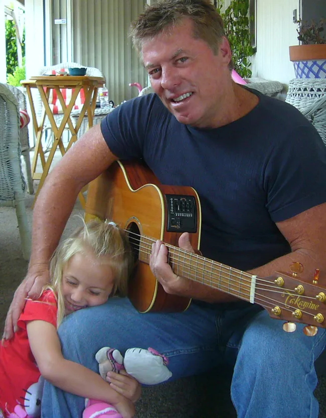 Holden Forrest playing an acoustic guitar on a porch while his young daughter hugs his knee
